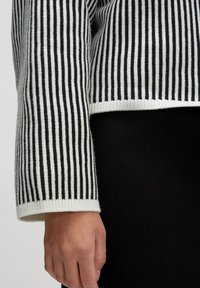 Gestreifter schwarzer und weißer Strickpullover mit gerippter Textur und weitem Bündchen. Der untere Rand ist mit einem durchgehenden weißen Band verarbeitet.