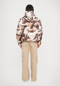 Giacca imbottita con stampa di orso geometrica in tonalità marrone e crema; presenta un cappuccio e una silhouette aderente. Abbinata a pantaloni cargo beige.