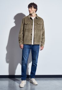 Olivová kordurová bunda se dvěma náprsními kapsami, v kombinaci s modrými džíny a béžovými teniskami, stojící proti bílé zdi.