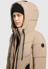 Beige vadderad jacka med en stor huva, svarta dragsko, och en texturerad yta. Har en svart cirkulär logga på vänster ärm.