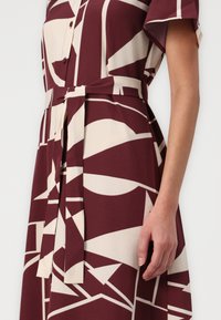 Robe à manches courtes en bordeaux foncé avec des motifs abstraits crème. Présente une ceinture à nouer et un système de fermeture à boutons, fabriquée à partir d'un tissu lisse.
