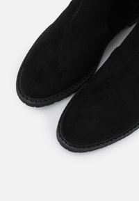 Botines de ante negro con textura suave, puntera afilada y suela de goma negra; detalle de costura a lo largo del borde.