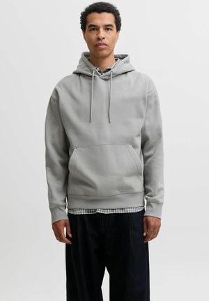 Homme debout portant un sweat à capuche gris clair par-dessus une chemise à carreaux et un pantalon noir ample, sur un fond clair uni.