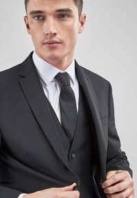 Fekete zakó sima textúrával, fehér inggel és textúrált fekete nyakkendővel párosítva. Egy gombos záródással rendelkezik.