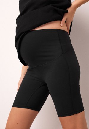 Kobieta w ciąży w czarnych rowerowych spodenkach ciążowych i czarnej bluzce, z jedną ręką opartą na dolnej części pleców.