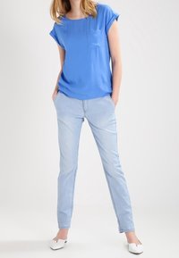 Modrá krátkosrstá halenka s přední kapsou je zkombinována se světle modrými úzkými džínami. Modelka má na sobě bílé ploché boty. Jednoduchý, ležérní outfit.