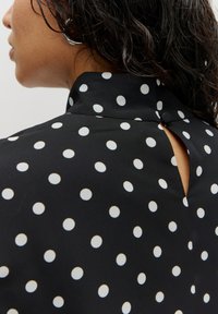 Schwarzes Hemd mit weißen Polka-Dots, ausgestattet mit einem Stehkragen und einem Schlüsselaugen-Detail im Rücken. Glatter Stoff mit leichtem Glanz.