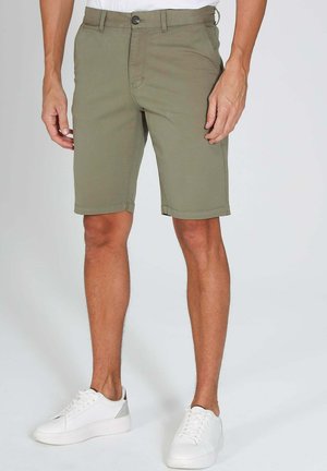 Homme portant un short olive jusqu'aux genoux et des baskets blanches, debout devant un fond clair uni.