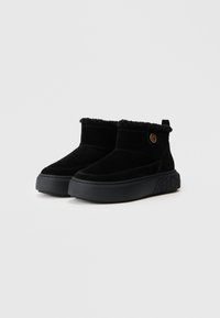Botas de tornozelo em camurça preta com um forro suave e felpudo, solas de borracha planas e um padrão em relevo discreto na sola. Acento de logotipo dourado na lateral.