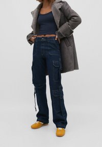 Pantalons cargo en denim bleu marine avec de multiples poches, taille haute et liens ajustables aux ourlets, associés à un manteau gris et des chaussures jaunes.