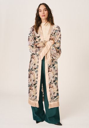 Femme debout portant une longue robe à motifs floraux avec des franges beiges sur un haut crème et un pantalon large bleu sarcelle, regardant droit devant.