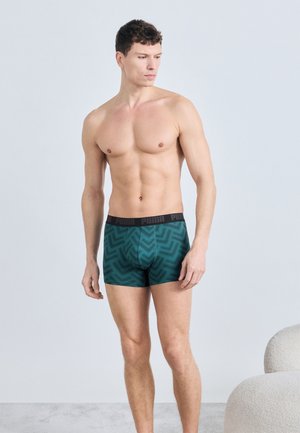 Άντρας χωρίς πουκάμισο, όρθιος, φοράει μπλε-πράσινα στυλιστικά ανδρικά εσώρουχα με μαύρη μέση Puma, κοιτάζοντας προς τα αριστερά του, με φόντο μια απλή ανοιχτή επιφάνεια.