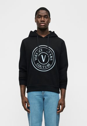 Črna kapuca z belim logotipom »Versace Jeans Couture«, ki jo nosi stojeca oseba, kombinirana s svetlo modrimi kavbojkami z znamčnimi bočnimi črtami.