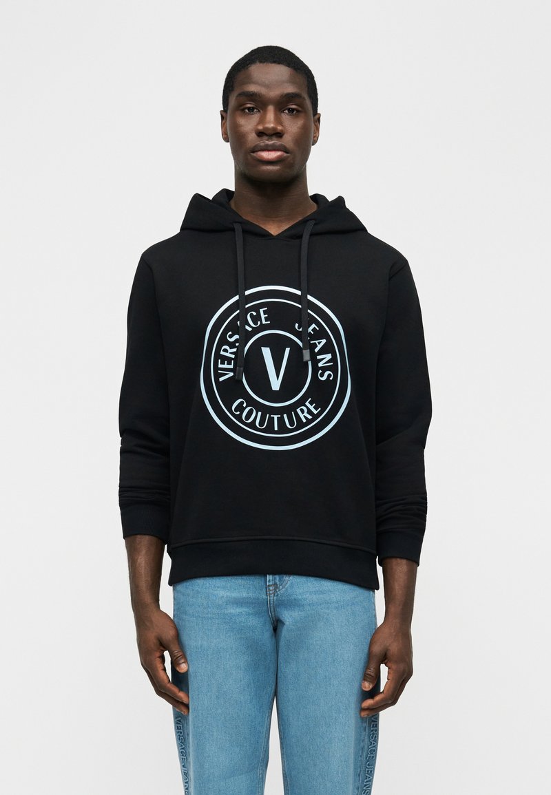 Μαύρο φούτερ με κουκούλα με λευκό λογότυπο «Versace Jeans Couture», που φοριέται από ένα άτομο που στέκεται, συνδυασμένο με ανοιχτό μπλε τζιν με ριγέ πλαϊνά λογότυπα.