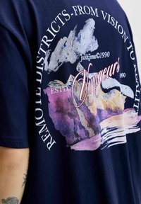 Granatowa bawełniana koszulka z kolorową abstrakcyjną grafiką i napisem. Wzór zawiera chmury, góry i słowo "Voyageurs".