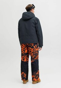 Juodas gobtuvas striukė, derinama su laisvai krentančiais oranžiniais ir juodais raštuotais slidinėjimo kelnais, turinčiais tekstūruotą medžiagą ir sutvirtintus rankogalius.