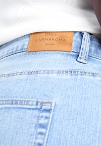 Svijetloplave traperice s smešnim kožnim oznakom "ONLY CARMKOMA Denim." Imaju šivane detalje i klasičan dizajn džepova.