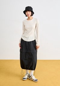 Frau trägt einen schwarzen Bucket Hat, ein beiges langärmliges Oberteil, einen schwarzen Midirock mit Zugbändern, weiße Socken und beige-navy Sneaker, steht drinnen.