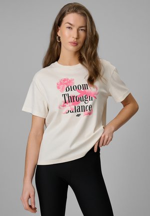 Kobieta o długich brązowych włosach, ubrana w białą koszulkę z różowym kwiatowym wzorem i napisem „Bloom Through Balance” oraz czarne spodnie.