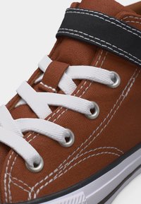 Καφέ πάνινο αθλητικό παπούτσι με λευκά κορδόνια, μαύρο ιμάντα με βέλκρο και λευκή ραφή, και λευκή λαστιχένια σόλα με μαύρο περίγραμμα.