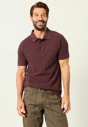 Lächelnder Mann mit Bart, der ein kastanienfarbenes kurzärmeliges Polohemd und olivgrüne Hosen vor einem einfarbigen Hintergrund trägt.