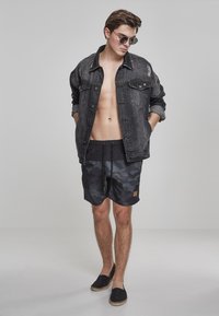 Veste en denim noir avec des détails usés, short de bain camouflage noir et espadrilles noires, complétés par des lunettes de soleil rondes.
