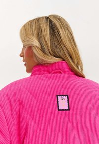 Różowa kurtka z korduroy o wysokim kołnierzu, z teksturowanej tkaniny, z prostokątną naszywką logo z tyłu, z tekstem i graficznymi detalami.