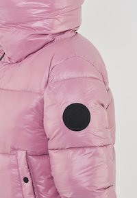 Giacca puffer rosa con finitura lucida, dotata di colletto alto e patch rotondo nero sul maniche sinistro. Include tasche frontali con bottone.