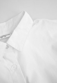 Biała koszula z kołnierzykiem i guzikami, z widocznymi guzikami oraz metką wewnątrz kołnierza pokazującą szczegóły rozmiaru.