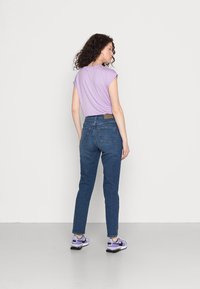 Top curto de manga curta lilás, jeans azul de cintura alta e ténis roxos. Cabelo encaracolado visível, de pé contra um fundo neutro.