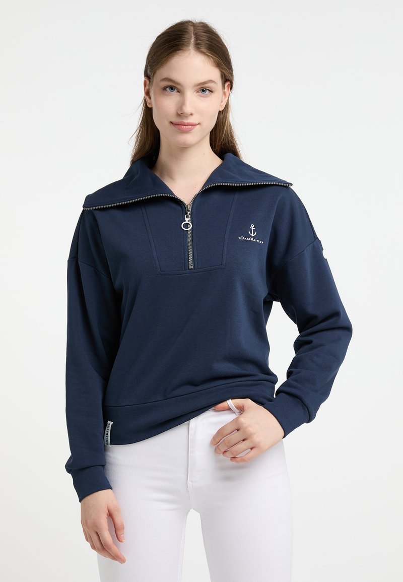 Jonge vrouw in een marineblauwe pullover met kwartrits en ankerlogo, met witte broek, staand tegen een plain witte achtergrond.