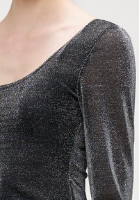 Svart langermet topp med scoop-neck, laget av et glitrende stoff som reflekterer lys, med en teksturert overflate med fint glitter.