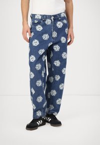 Blue denim pants featuring a repeated dice pattern in white. Loose fit with five-pocket design and a button closure. Black sneakers complement the look.