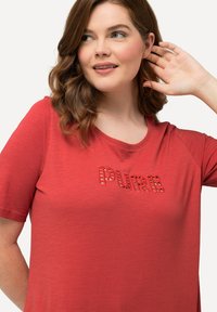 Camiseta roja de manga corta con escote redondo. Texto "PURE" en diseño de recortes en el pecho. Tejido suave con una textura lisa.