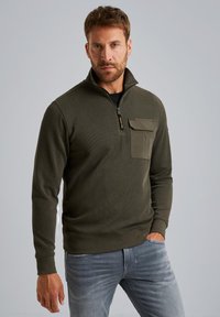 Sudadera de color verde oliva con cremallera, hecha de tejido texturizado, con un bolsillo en el lado izquierdo del pecho, combinada con vaqueros de color azul claro.
