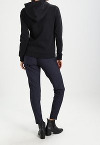 Felpa nera con cappuccio dal design aderente, abbinata a pantaloni a righe verticali blu navy. La modella indossa stivaletti neri alla caviglia.