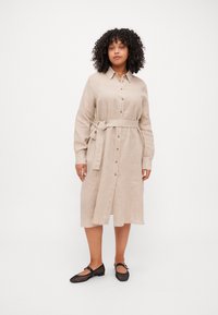 Abito camicia in lino beige con colletto, bottoni frontali e cintura legata in vita. Le ballerine in rete nera completano l'outfit.