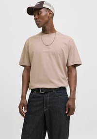 Beige katoenen t-shirt met korte mouwen en geborduurd logo op de borst, gecombineerd met donkere spijkerbroek met contrasterende stiksels en riem.