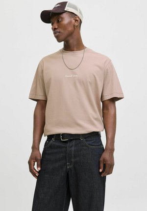 Beige Baumwoll-T-Shirt mit kurzen Ärmeln und gesticktem Logo auf der Brust, kombiniert mit dunklen Jeans aus Denim mit kontrastierenden Nähten und Gürtel.