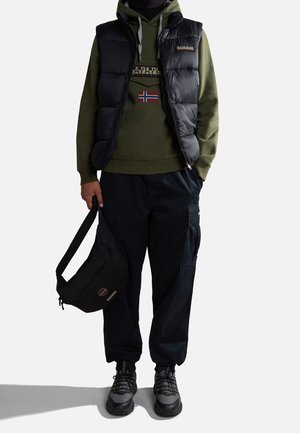Person wearing black puffer vest over olive hoodie with flag patch, black cargo pants, gray sneakers, holding black sling bag.