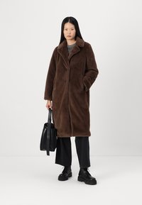 Cappotto di pelo finto marrone con colletto, chiusura a bottone e tasche laterali. Indossato sopra un maglione a trama e pantaloni neri, abbinato a stivali e borsa.