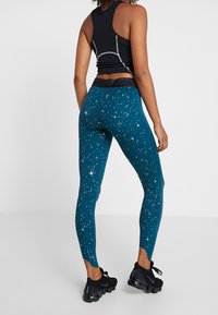 Leggings ceñidos color teal con un patrón estelar cósmico; textura suave, cintura alta, combinados con zapatillas deportivas negras.