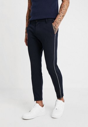 Uomo con pantaloni aderenti blu navy con strisce laterali bianche, mani nelle tasche, abbinati a sneaker bianche e un top senza maniche blu navy lavorato a trama.