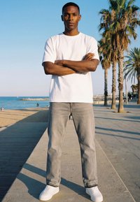 Uomo con t-shirt bianca e jeans grigi, braccia incrociate, in piedi su una passeggiata soleggiata sul lungomare con palme e oceano sullo sfondo.