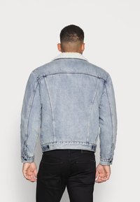 Ljusblå denimjacka med en fluffig vit pälskrage, synliga stickningdetaljer och en något snäv passform. Bakifrån syns två vertikala sömmar.