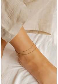 Cavigliera in tonalità oro composta da due catene delicate, una liscia e una con un design di perline, indossata su una caviglia nuda.