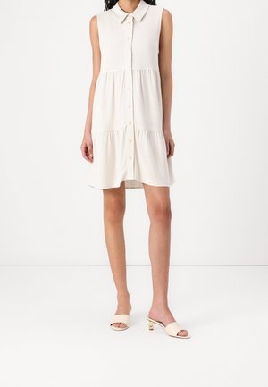 Kobieta w bezrękawnikowej beżowej sukience zapinanej na guziki z kołnierzykiem oraz beżowych sandałach na niskim obcasie z odkrytymi palcami, stojąca na białej podłodze na tle białej ściany.