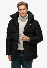 Jonge man in een zwarte pufferjack over een beige turtleneck sweater en blauwe spijkerbroek, die met één hand de rits van de jack aanpast.