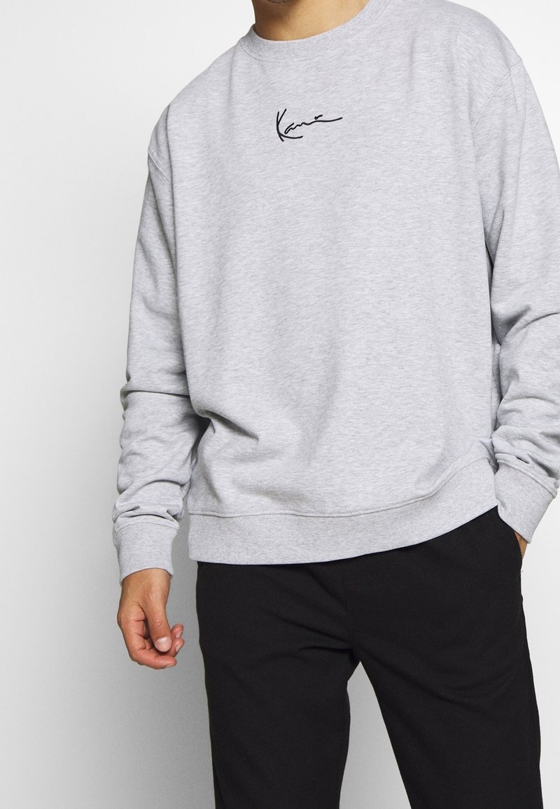 Homme portant un sweat-shirt gris clair à manches longues et col rond avec un logo noir style signature sur la poitrine et un pantalon noir, mains détendues.
