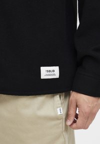 Black fleece pullover with a white woven label on the hem, featuring text and branding. Paired with light beige pants and a smaller label visible.